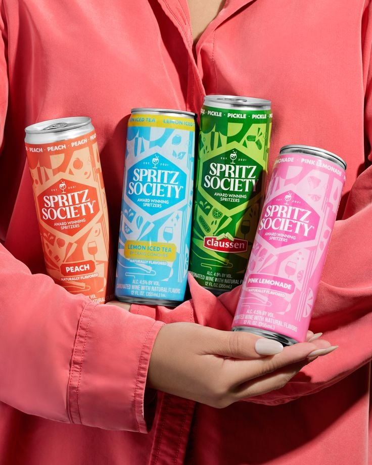 Vibrant Canned Beverage Showcase — A lifestyle product photograph featuring four slim canned spritz beverages held together in both hands against a soft coral-c